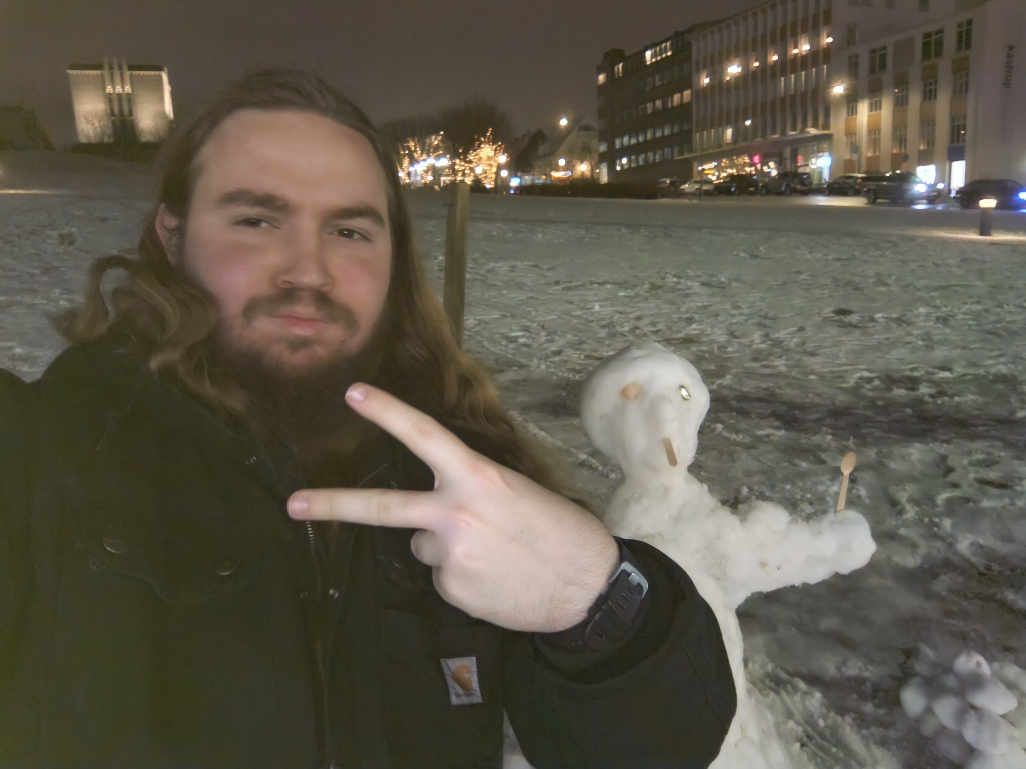 A person with long hair and a beard takes a selfie at night in a snowy outdoor area, holding up a peace sign. Behind them is a small snow figure with simple features made from sticks and small objects. In the background, buildings and trees are lit with warm lights, and parked cars line the street.
