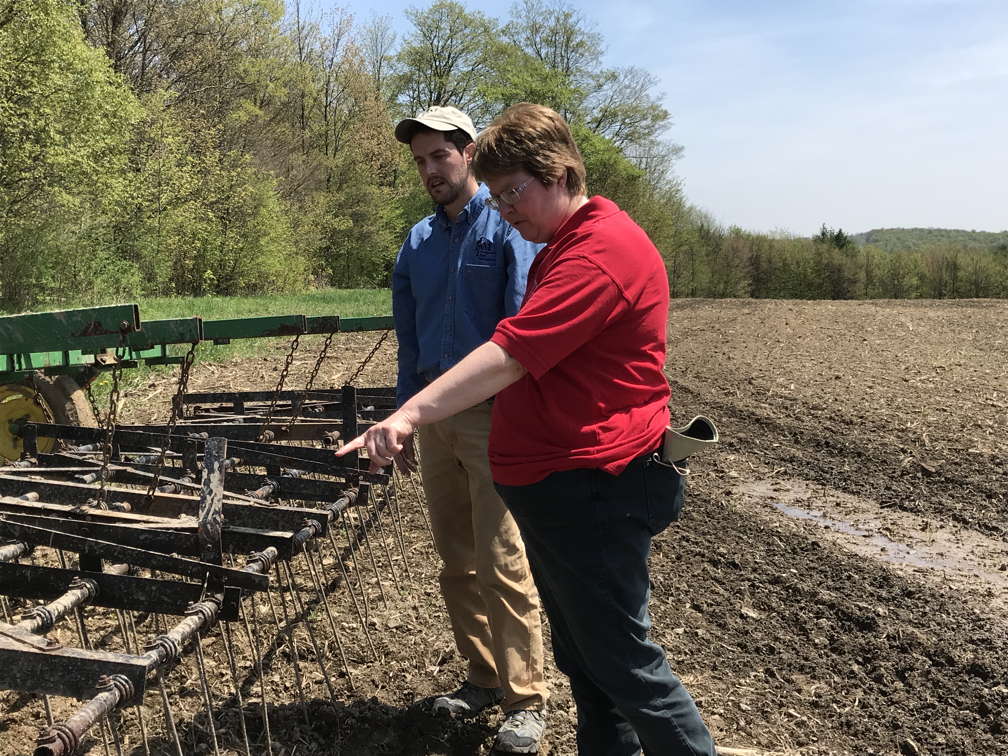 A woman in a red shirt pointing at farm equipment with a man looking on.