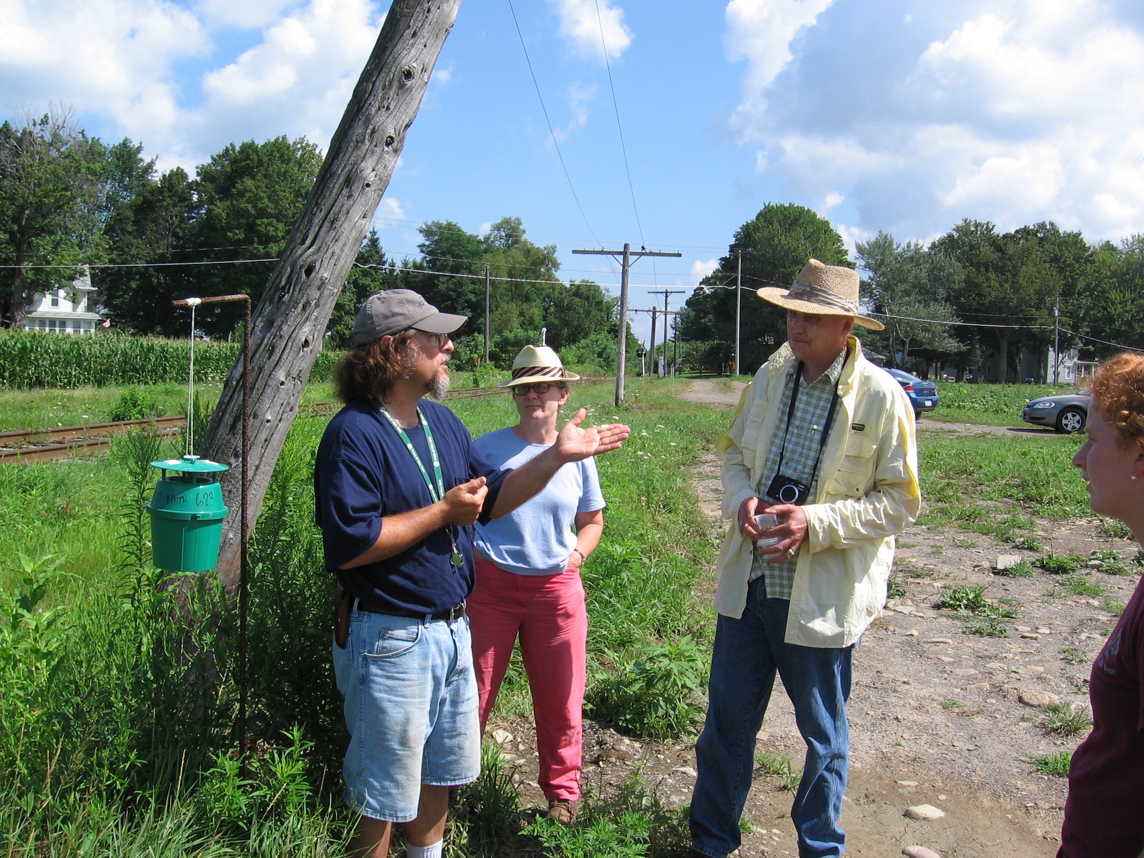 A man motions with his hand to three other people listening to him. He is standing along a corn field edge with a pheromone trap on a pole behind him.