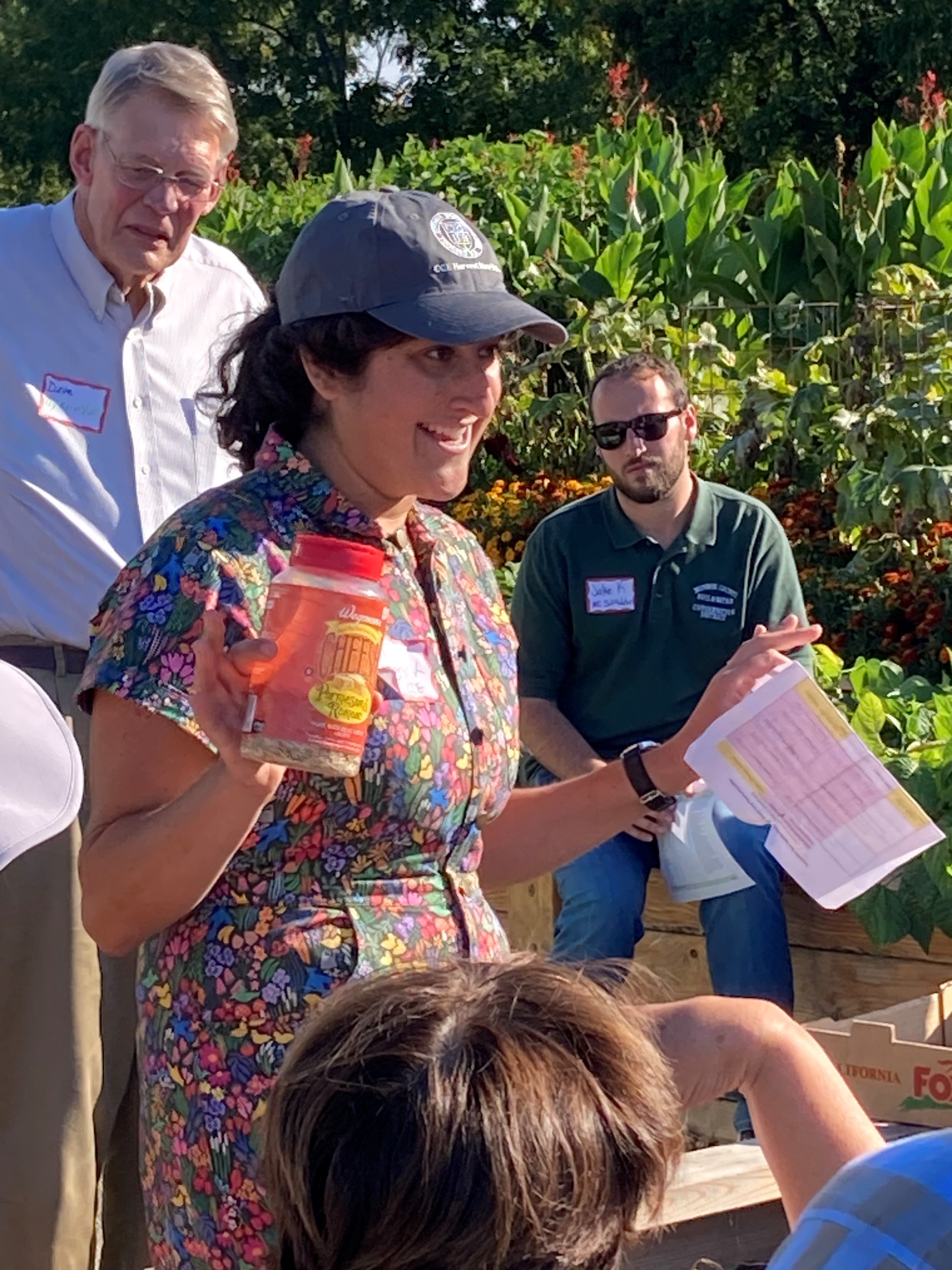 A woman holds up a repurposed grated cheese shaker while speaking in an urban garden during a meeting.
