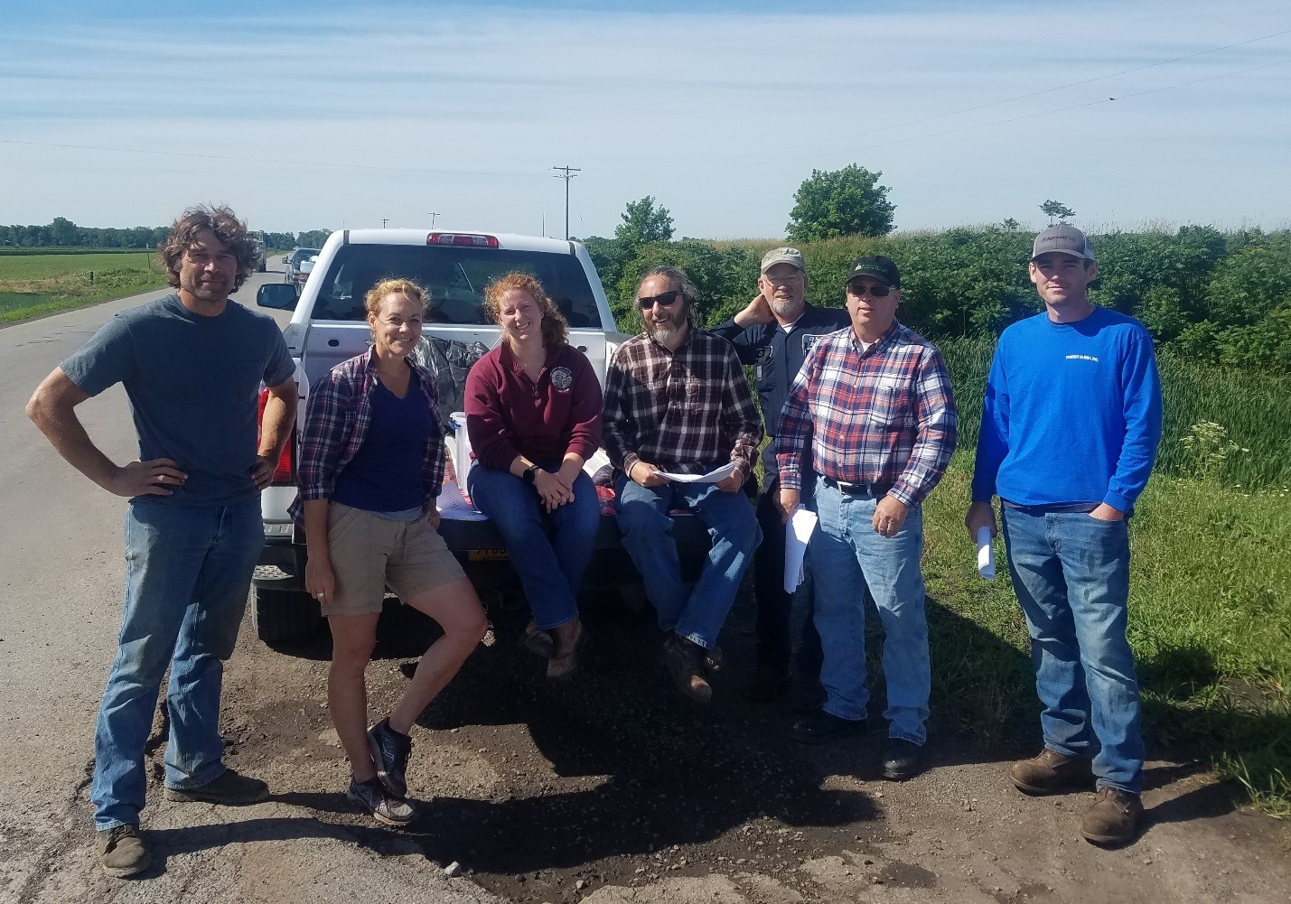 Seven people pose for a photo along the side of a road in the Elba muck. A couple of them are sitting on the tailgate of a white pickup truck.