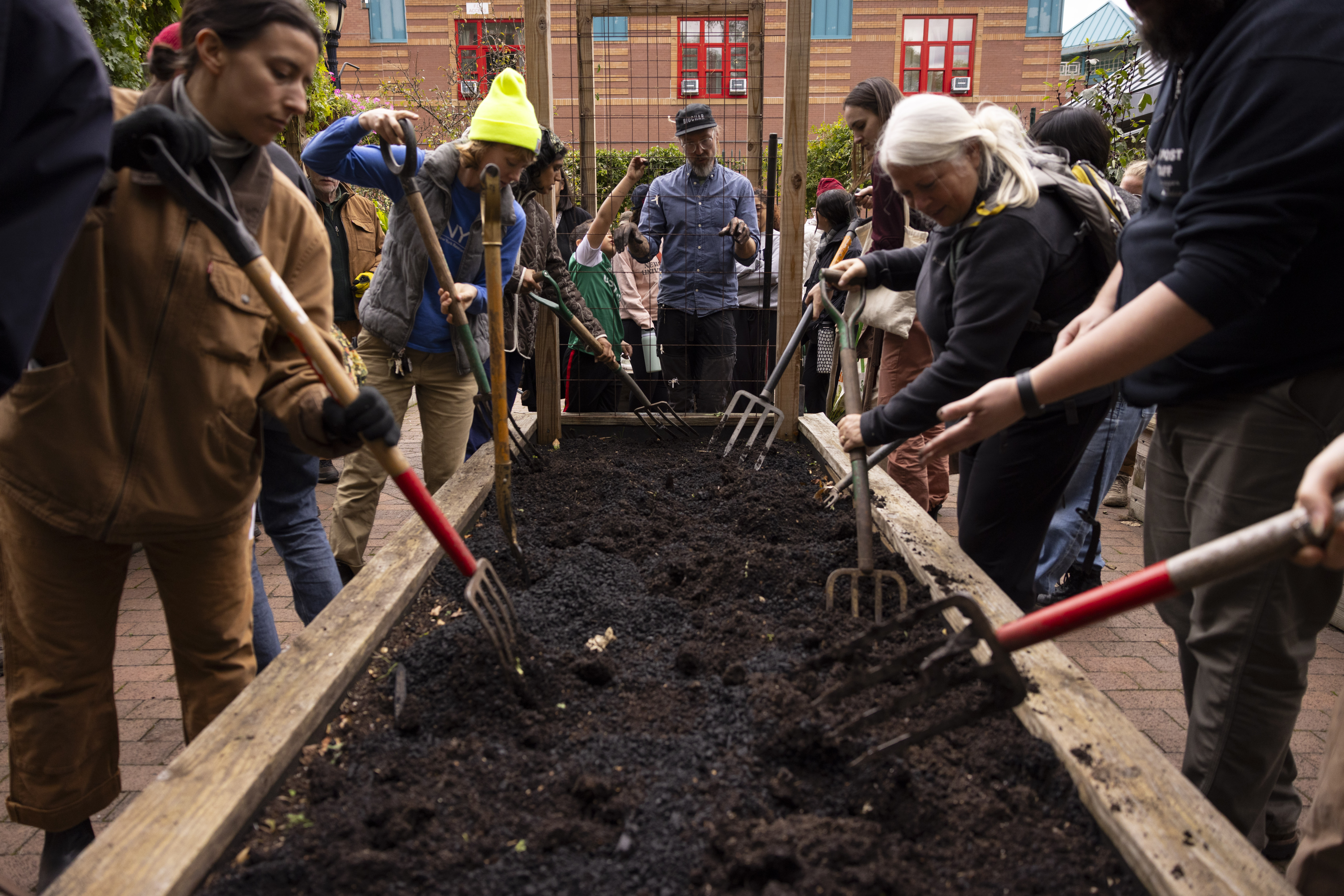 Several people surround a raised planting bed using garden forks to incorporate biochar into the soil.