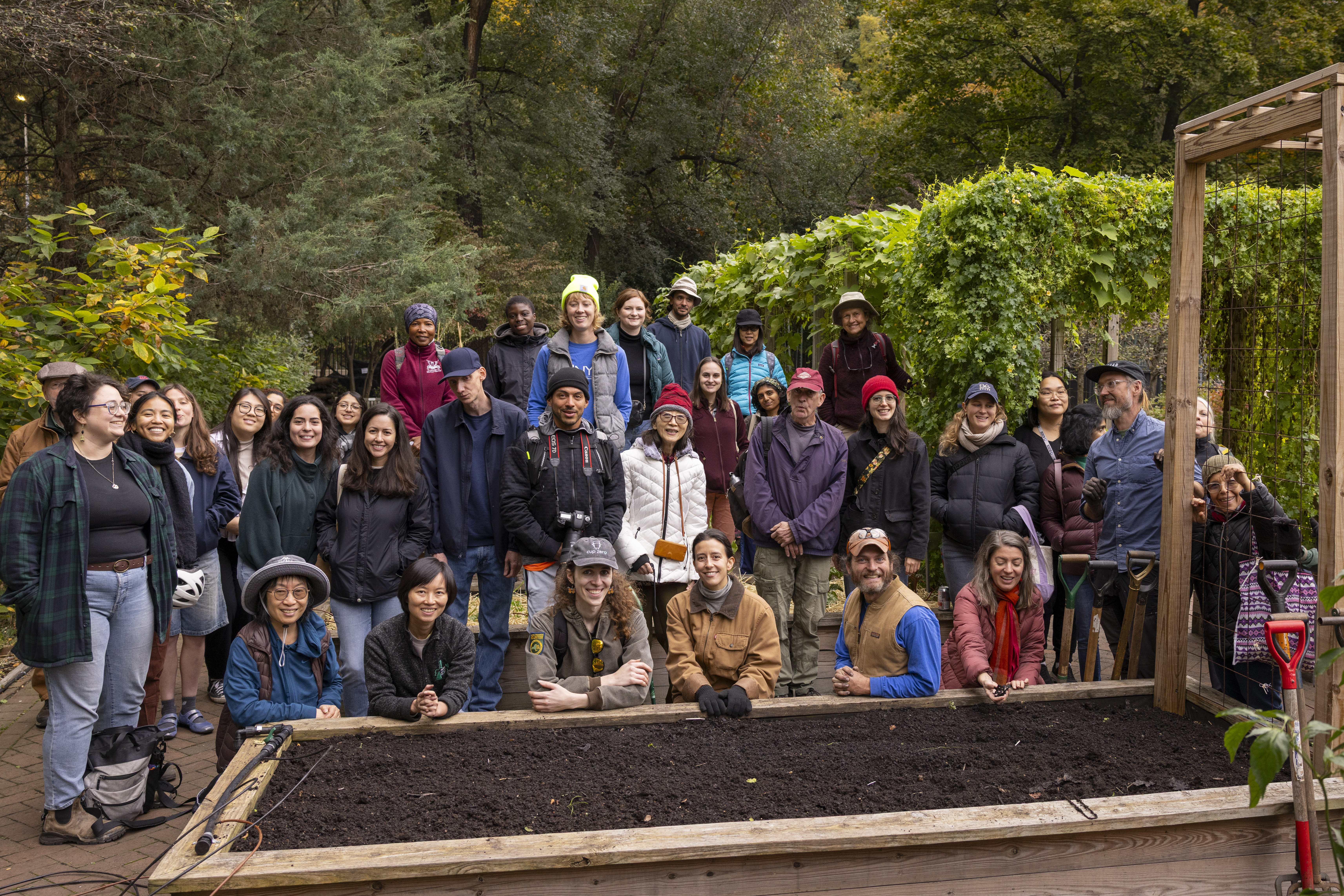 A large group of people pose together outdoors behind a raised garden bed filled with soil. They are smiling and dressed warmly, surrounded by trees and garden structures
