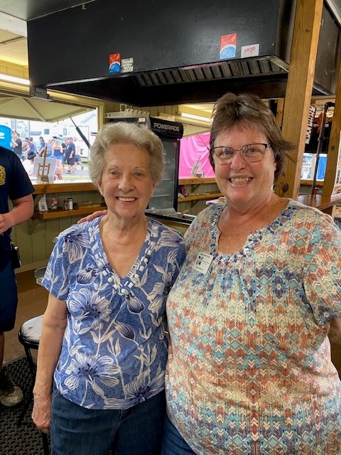 Two women standing side by side indoors, smiling at the camera. The woman on the left has short gray hair and is wearing a blue floral patterned shirt and dark jeans. The woman on the right is wearing glasses and a multicolored patterned blouse with a name tag pinned to it. They are standing in what appears to be a kitchen or food service area, with a black ventilation hood above them, wooden beams, and a glass window behind them showing people outside.