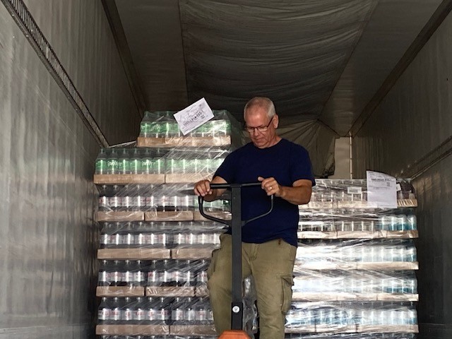 A man standing inside a large trailer, holding the handle of a manual pallet jack. Behind him are multiple stacked pallets wrapped in plastic, filled with cases of canned goods or packaged items. The interior of the trailer is lined with metal walls and a fabric ceiling, and paperwork is attached to one of the wrapped pallets.