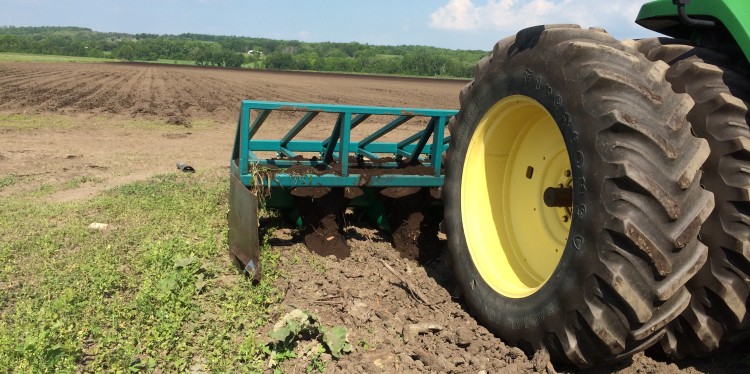 A closeup of a tractor wheel and a plowing implement. In the background is a freshly plowed field.