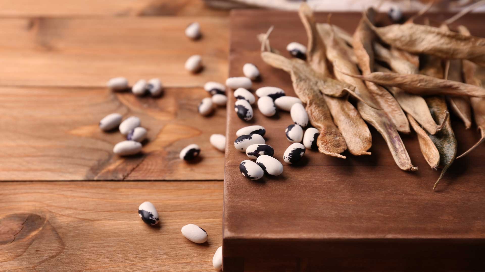 Dried bean pods and black-and-white beans scattered across a wooden table and cutting board. The image has warm brown tones and a rustic farm-style feel, with several loose beans spread in the foreground and dried pods piled on the right side.