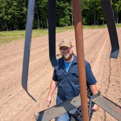 A smiling man wearing glasses, a beard, and a tan baseball cap stands in a freshly tilled farm field. He is holding a black plastic seedling tray while looking up toward the camera. Large metal farm equipment parts hang in the foreground, partially framing him. Behind him are neat rows of soil and a line of green trees under a bright sunny sky.