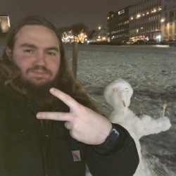 A person with long hair and a beard takes a selfie at night in a snowy outdoor area, holding up a peace sign. Behind them is a small snow figure with simple features made from sticks and small objects. In the background, buildings and trees are lit with warm lights, and parked cars line the street.