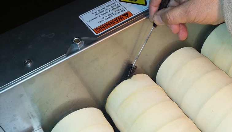 A hand holds a small bottle brush, using it to clean the side wall along sets of absorber donuts with a brush washer conveyor produce washing system.