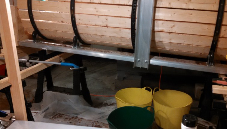 Lower view of the metal support frame holding a root barrel washer. There is debris on the frame and the floor below. Two yellow wash buckets and one green one sit on the floor below the machine.