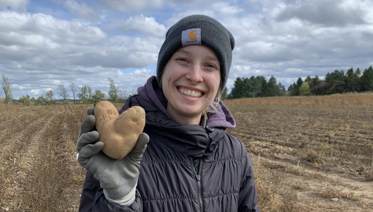 A smiling woman in a dark jacket and beanie cap holds up a heart shaped potato. She is standing in front of a potato field.