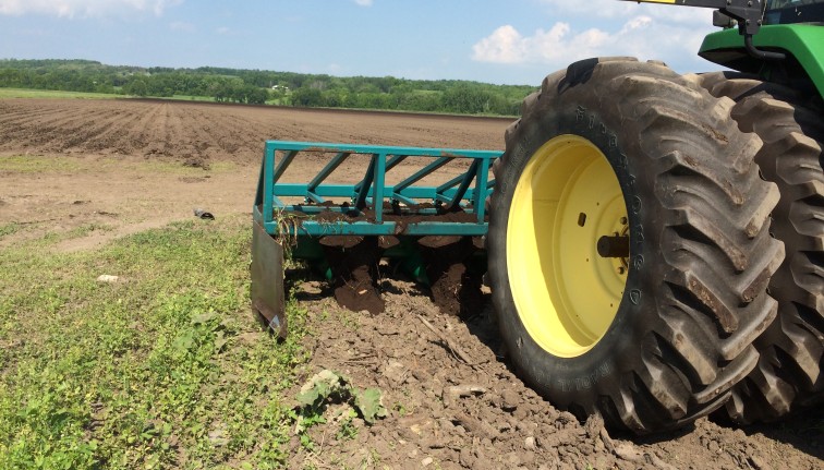 A closeup of a tractor wheel and a plowing implement. In the background is a freshly plowed field.