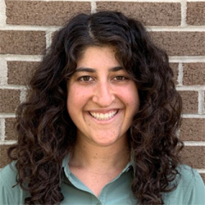 Headshot of Lori Koenick: Smiling woman in sage colored shirt, standing in front of a brick wall.