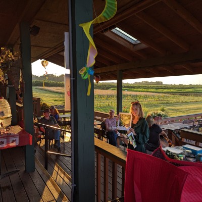 Several people on a covered deck at sunset in a country setting. A woman holds up a handout.