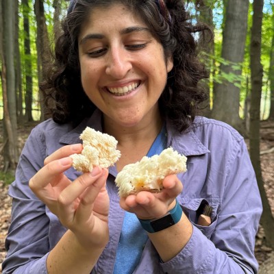 A woman standing in a forest is holding mushrooms in her hands. She is looking at them and smiling.