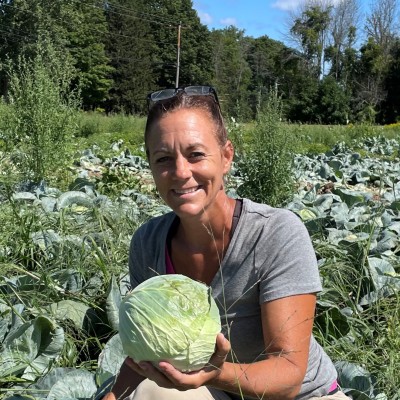 A woman squatting down in a field of cabbage, holding a head in her left hand.