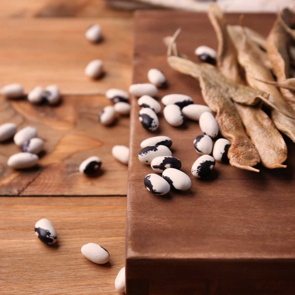 Dried bean pods and black-and-white beans scattered across a wooden table and cutting board. The image has warm brown tones and a rustic farm-style feel, with several loose beans spread in the foreground and dried pods piled on the right side.