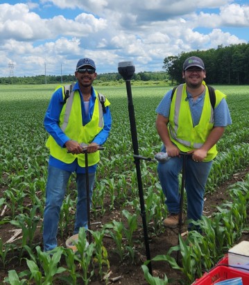 Two men stand in a field with sampling equipment.