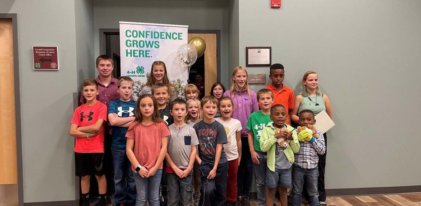 A group of children and teenagers are standing and smiling in a well-lit indoor hallway. They are arranged in rows, with some taller individuals towards the back. Behind them, is a large green and white banner that reads "CONFIDENCE GROWS HERE." The 4-H clover logo is prominently displayed on the banner.