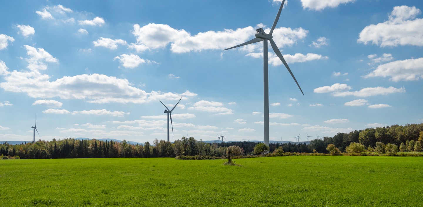  A vibrant, wide-angle shot of a green field under a dynamic blue sky with scattered clouds. In the foreground, a large wind turbine with three blades stands prominently. In the distance, other wind turbines are visible across a line of trees.