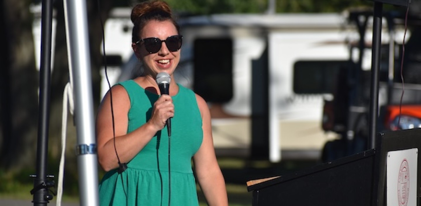 A woman wearing a green dress and sunglasses speaks into a microphone at an outdoor event, standing at a podium with the Cornell Cooperative Extension logo