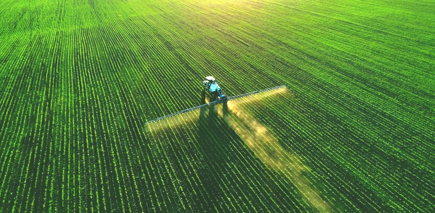 Aerial view of a tractor spraying crops across a large green agricultural field, with sunlight casting long shadows and highlighting the neat rows of plants.