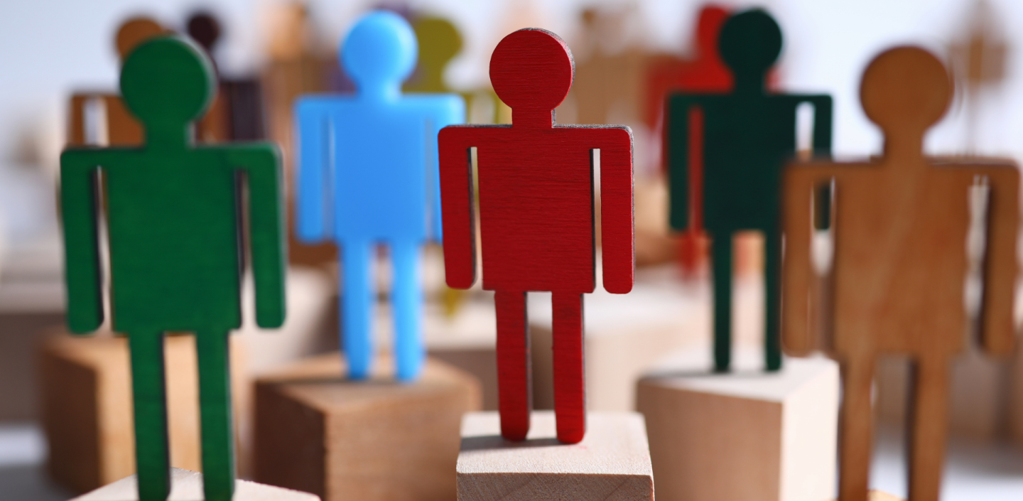 A group of various colored wooden and plastic stylized human figures standing on individual wooden blocks, with a red figure in the foreground.