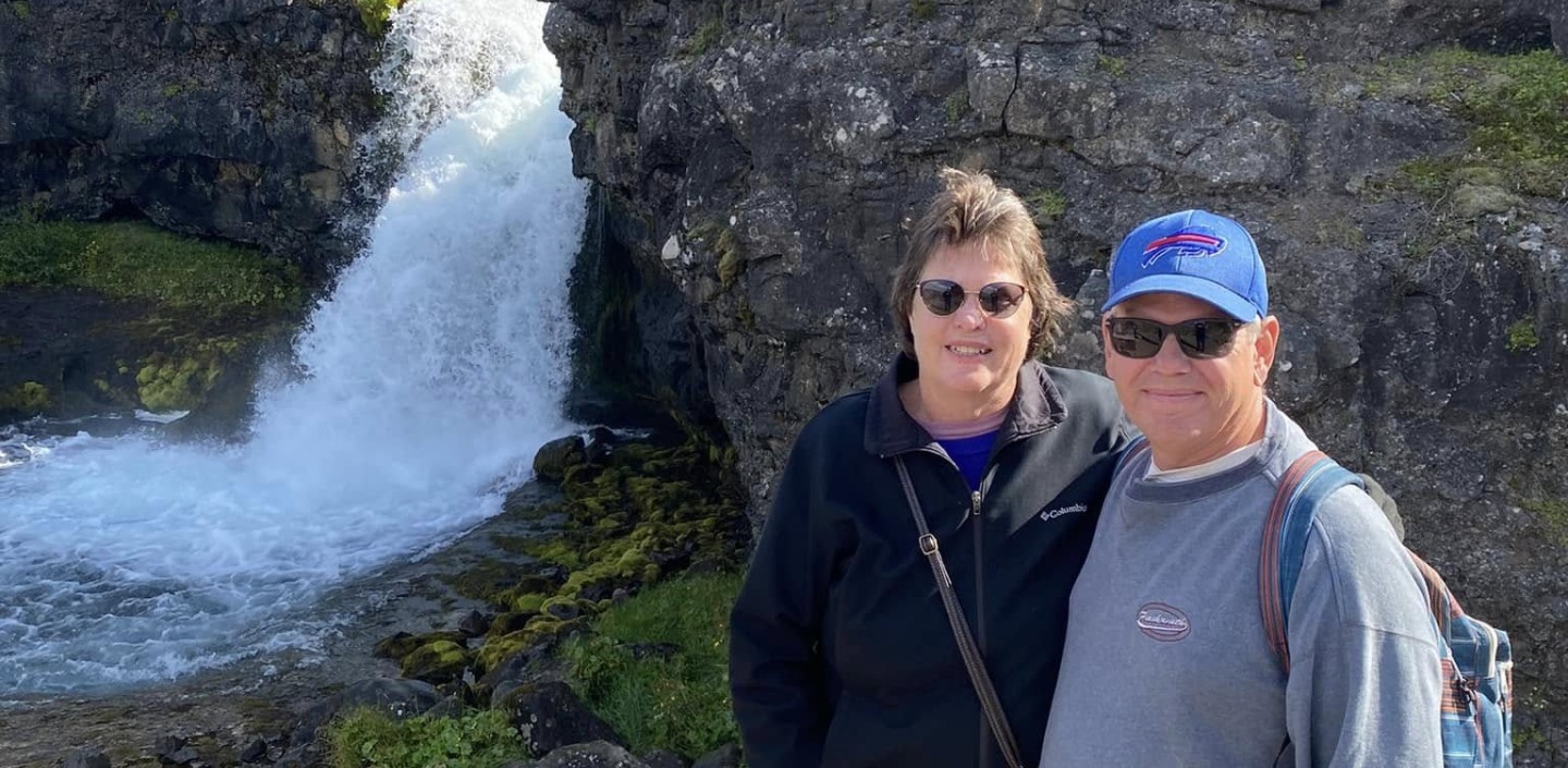 Two adults standing together on a rocky overlook in front of a small waterfall cascading between rugged cliffs. The woman is wearing sunglasses, a black jacket, and jeans. The man is wearing sunglasses, a blue baseball cap, a gray sweatshirt, jeans, and a backpack. Moss-covered rocks and flowing water are visible below, with steep rocky hills rising in the background under a clear blue sky.