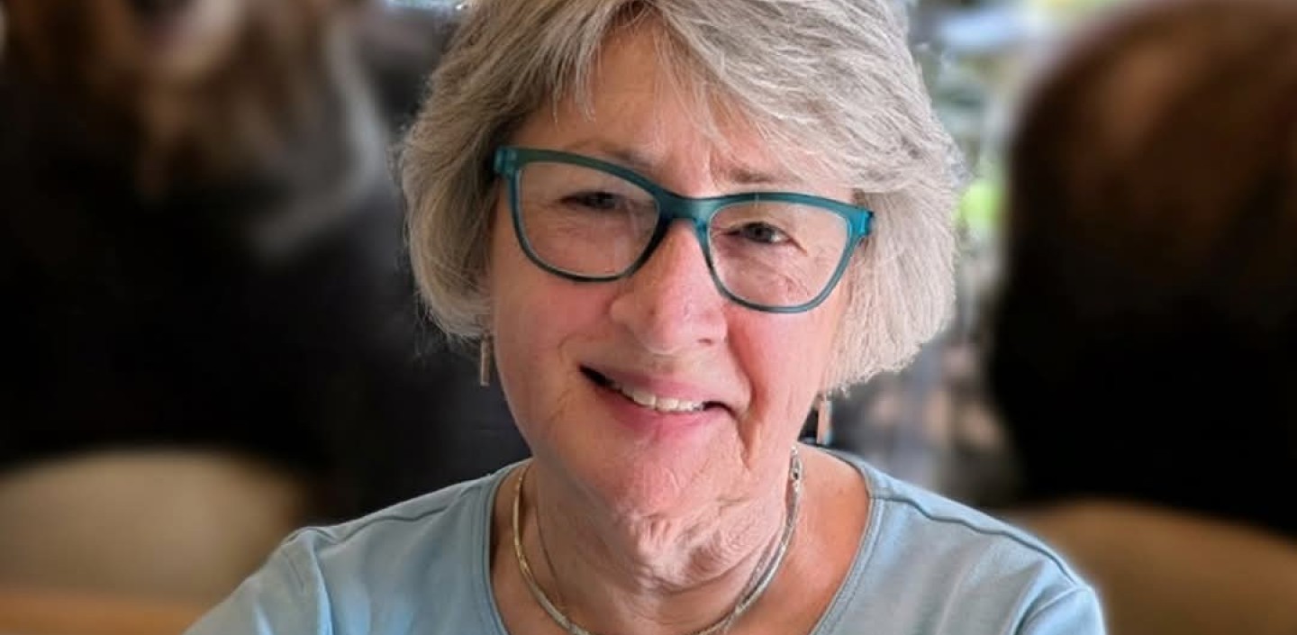 An older woman with short gray hair and teal glasses smiles at the camera while seated indoors. She is wearing a light blue long sleeve shirt and layered turquoise jewelry. The background is softly blurred, showing large nature themed artwork and two bears sitting behind her.