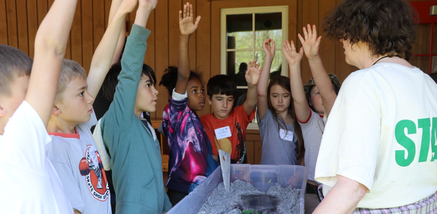 A group of children raising their hands around a homemade stream table as the instructor explains the activity.