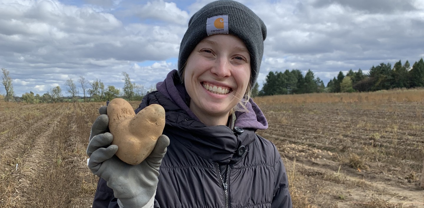 A smiling woman in a dark jacket and beanie cap holds up a heart shaped potato. She is standing in front of a potato field.