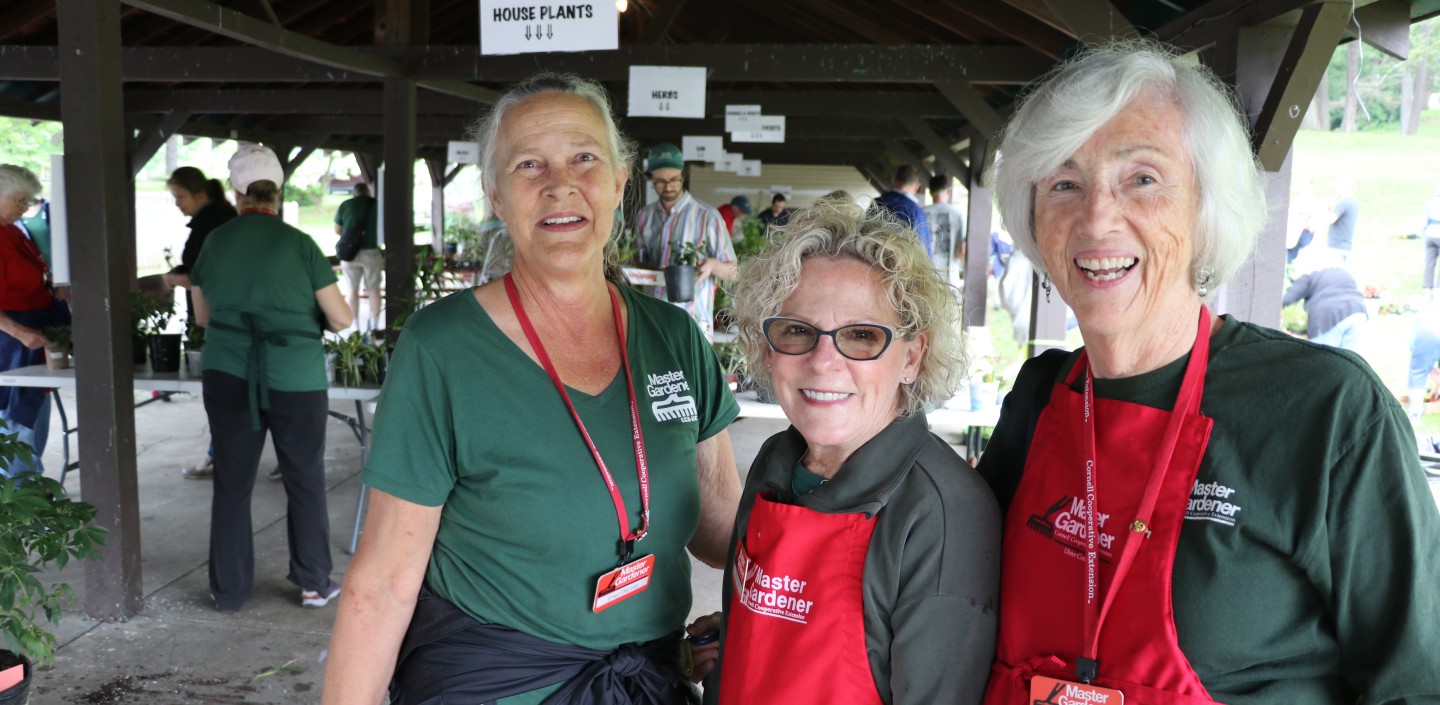 Three CCE Ulster Master Gardeners stand and smile in aprons and green Master Gardener shirts.