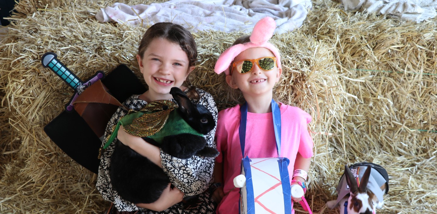 2 4-H'ers pose with their rabbits. On the left, the girl is holding a black rabbit, wearing a black dress, with a fake sword on her back. The other girl, on the right, is dressed in all pink with a drum and bunny ears, like the Energizer Bunny, while her black and white rabbit sits to her left.