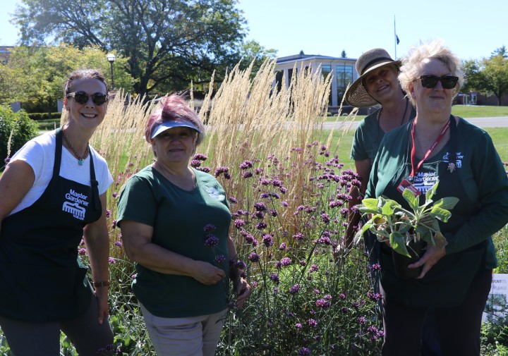 4 Master Gardeners stand holding various plants, wearing Master Gardener shirts and aprons, in the demonstration garden.