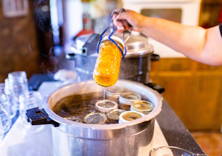 A large canner of water on a stove with canning jars in it. One jar is being removed from the pot using jar tongs.