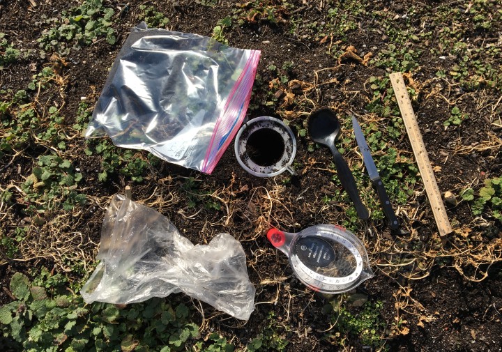 Several bulk density soil sampling tools laying in a group on the ground. There is a plastic baggie, a large spoon, a knife, ruler, and measuring cups.
