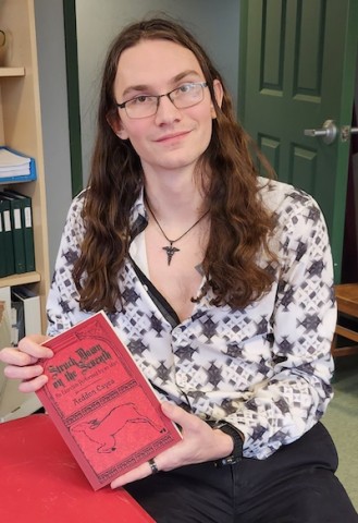 Person with long brown hair and glasses sitting indoors, holding a red book titled 'Stark Raven on the Lam: The Adventures of Addison Caven.' They wear a black and white patterned shirt and a pendant necklace. Behind them is a green door and a bookshelf with binders and books; a red table is in the foreground.