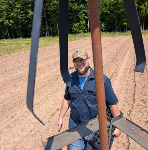 A smiling man wearing glasses, a beard, and a tan baseball cap stands in a freshly tilled farm field. He is holding a black plastic seedling tray while looking up toward the camera. Large metal farm equipment parts hang in the foreground, partially framing him. Behind him are neat rows of soil and a line of green trees under a bright sunny sky.