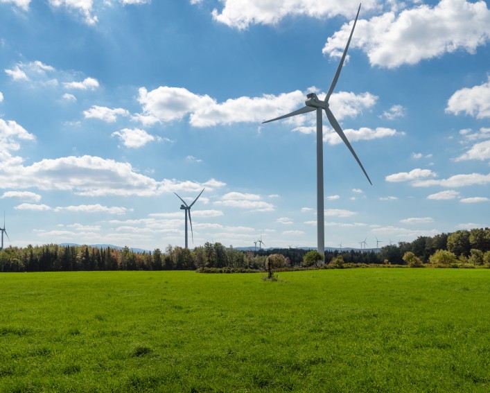  A vibrant, wide-angle shot of a green field under a dynamic blue sky with scattered clouds. In the foreground, a large wind turbine with three blades stands prominently. In the distance, other wind turbines are visible across a line of trees.