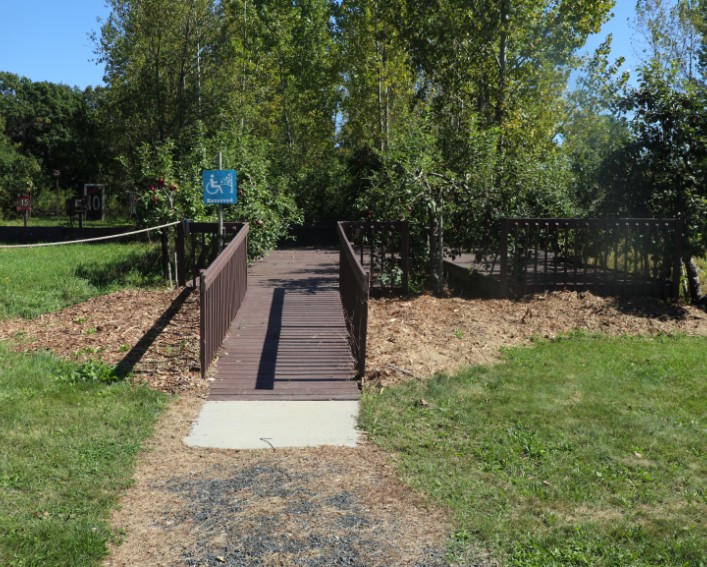 A wide brown wooden ramp with railings on the side lead up to a wooden raised platform around apple trees. There is a blue handicap accessible sign along the ramp.