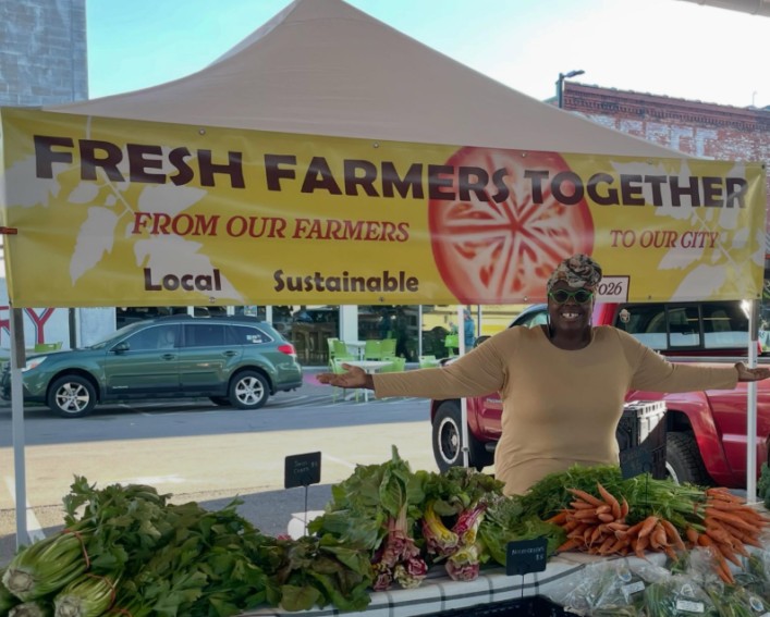 A woman stands behind a table of produce smiling with her arms held open as if to draw attention to the produce. Behind her is a bright yellow banner that says, " Fresh Farmers Together from our farmers to our city, local, sustainable" with an image of a tomato cut in half.