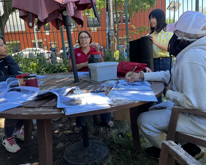 A woman in a red polo shirt is seated at an outdoor table and talking. Three people, two sitting and one standing, are listening to her. There are packets of information and notebooks on the table.