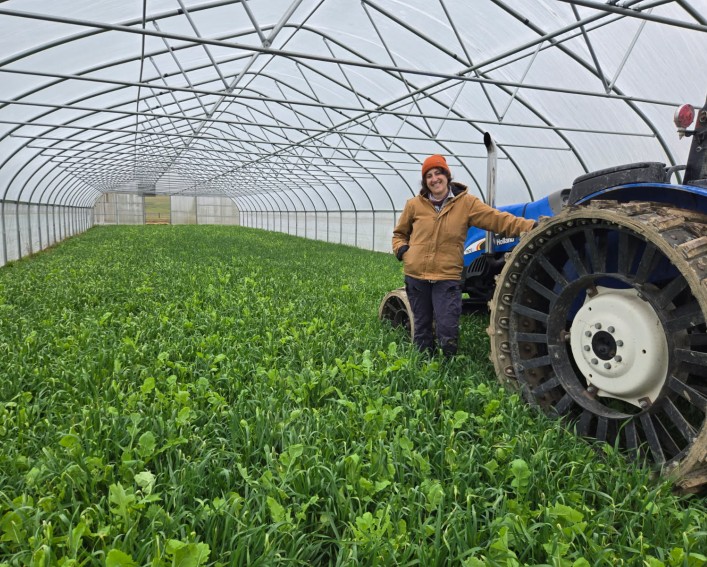 A woman leans with one hand on a tractor within a high tunnel. She is wearing a heavy coat and an orange hat. There are cover crops at her feet filling the high tunnel.