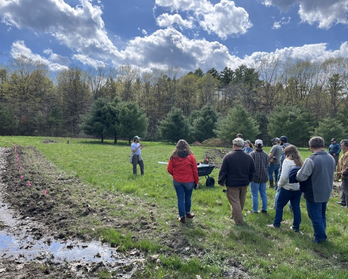 About a dozen people standing outside listening to a person talking to them about climate mitigation steps to take on their farms.