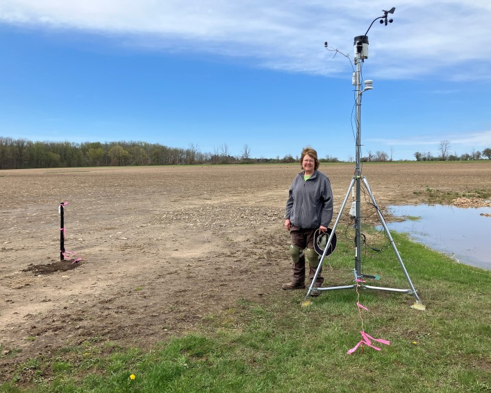 A woman stands near a weather station along an unplanted field.