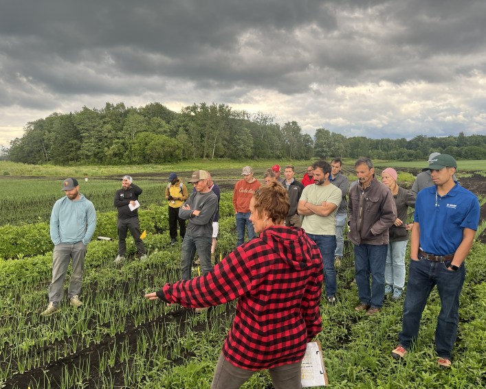 A woman in a red and black checkered jacket shows her field trial plots to a dozen onion growers. They are standing in a muck onion field.