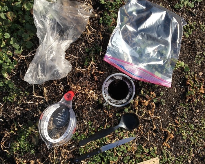 Supplies laying on the ground in preparation for taking a bulk density sample: two plastic bags, a measuring cup, a ruler, a kitchen knife and long spoon, and a plastic ring held in place with two landscape staples.
