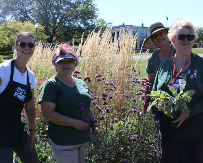 4 Master Gardeners stand holding various plants, wearing Master Gardener shirts and aprons, in the demonstration garden.