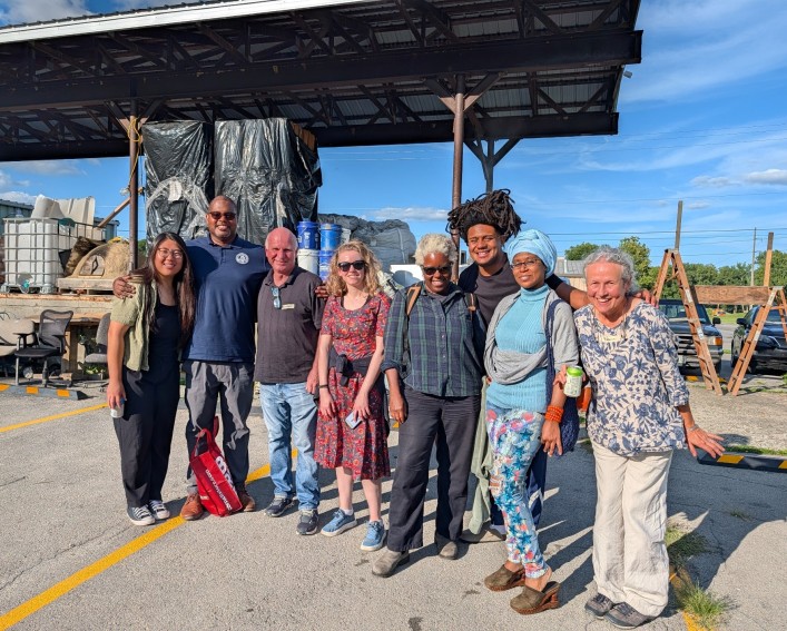 Eight people pose in a line outside in an industrial parking area setting. They are all smiling.